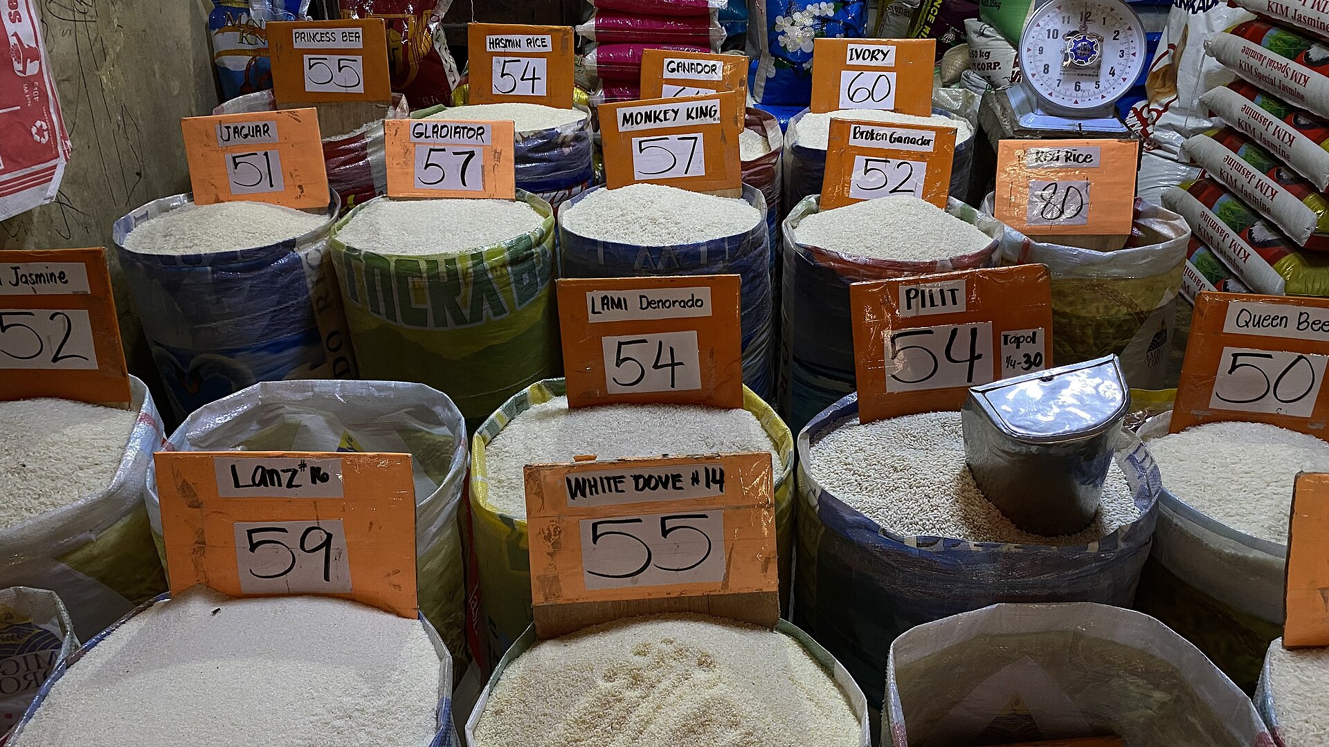 Open sacks of rice at a market stall, each with an orange handwritten price-per-kilo sign from 50 to 80 pesos